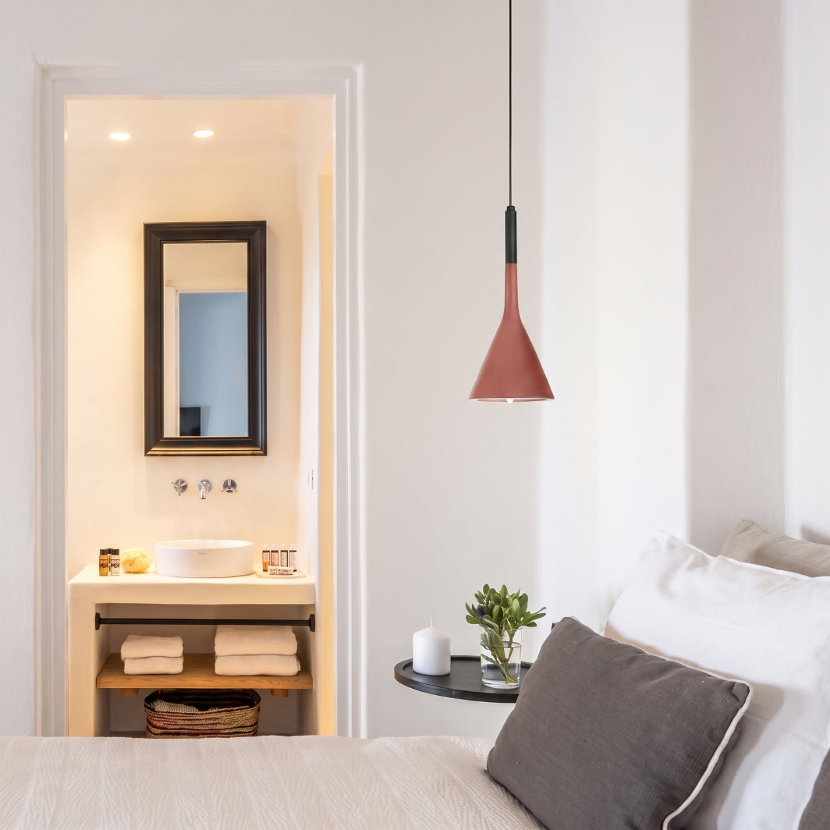 A modern bedroom scene featuring a distinctive conical pendant light in terracotta red with a black top accent, hanging over a bed with white pillows and a gray cushion. An open doorway in the background reveals a minimalist bathroom with a white vessel sink and a dark wood mirror.