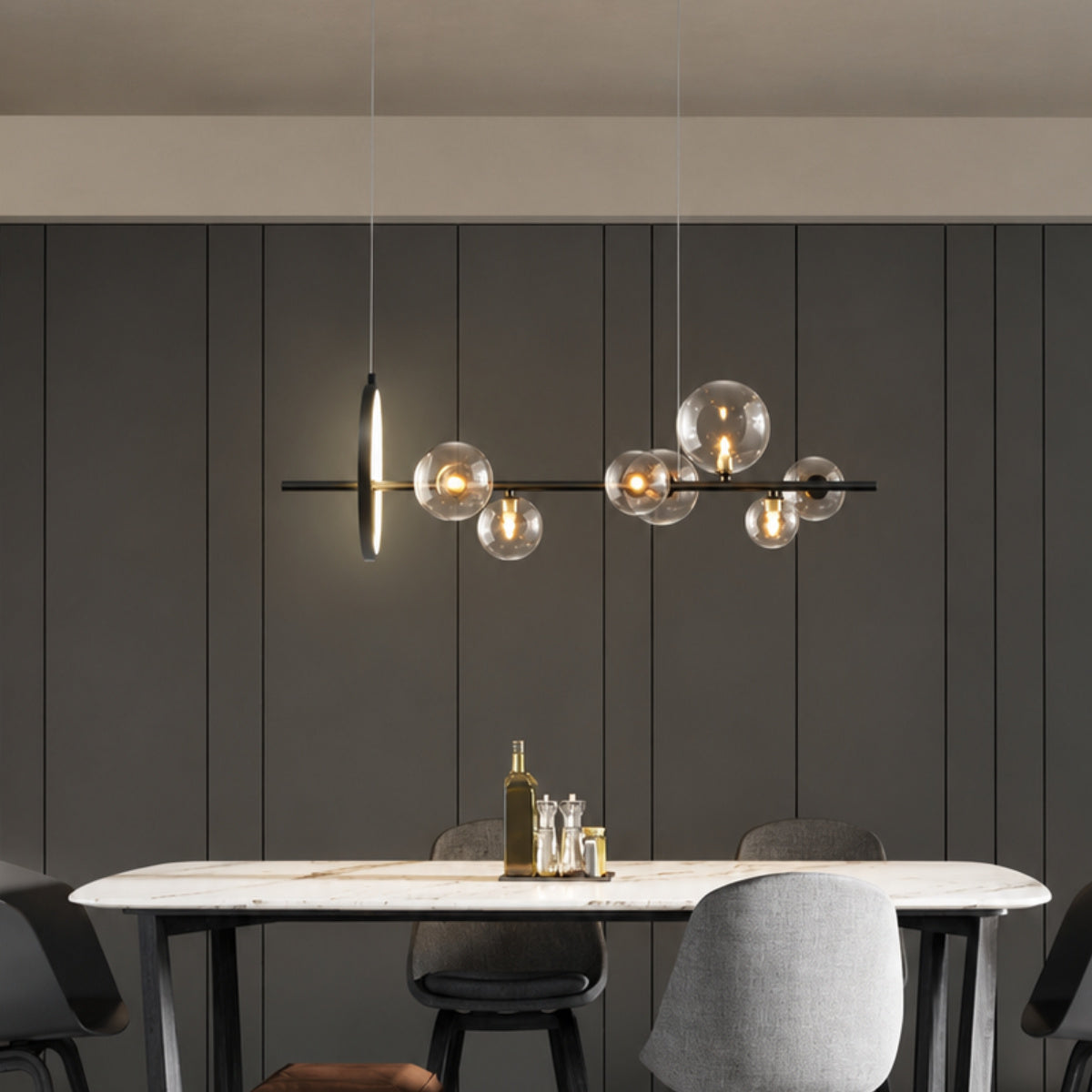 Modern linear pendant light with multiple clear glass globes and warm exposed bulbs, hanging above a dining table in a contemporary interior with dark paneled walls.