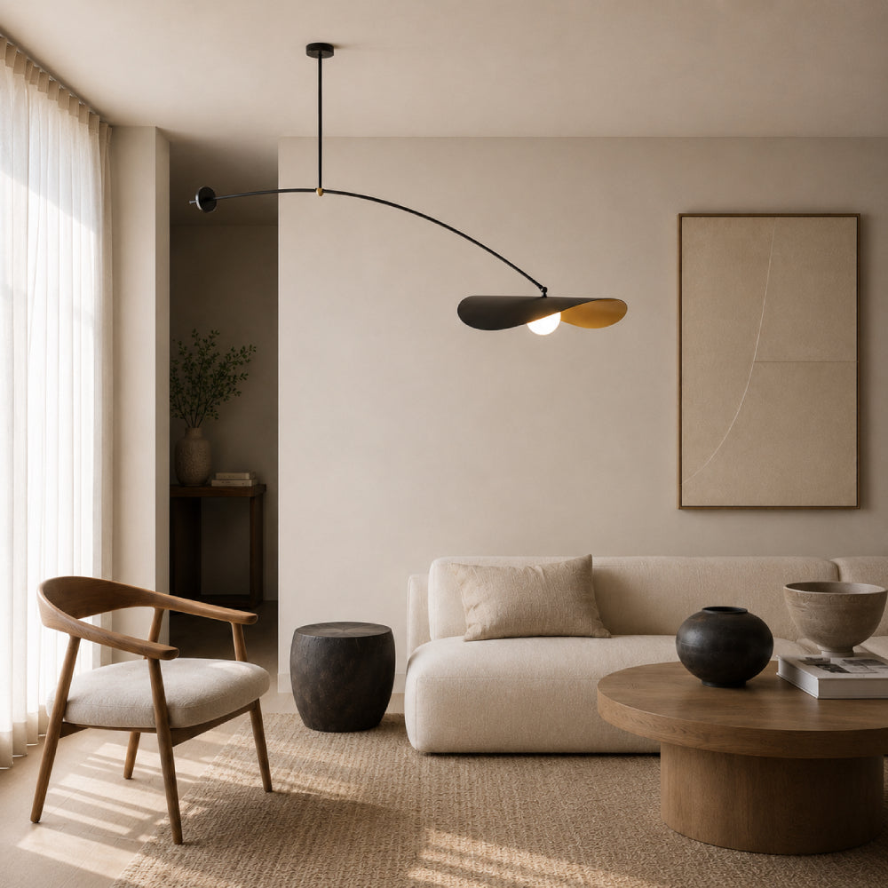 A modern living room with a sculptural black metal pendant light featuring two overlapping shades, one matte black and one warm gold. The room has beige walls, a cream sofa, a wooden armchair, and a round coffee table.
