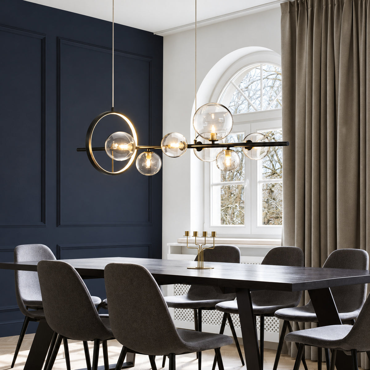 Modern brass and black linear pendant chandelier with clear glass bubble globes hanging over a dark wood dining table with gray upholstered chairs in a stylishly decorated room.